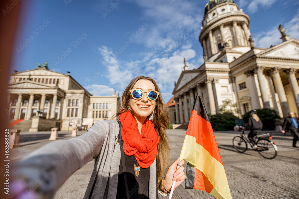 Fototapeta premium Młodej kobiety selfie turystyczna robi fotografia z niemiec flaga na Francuskim kościół i opery tle w Berlin
