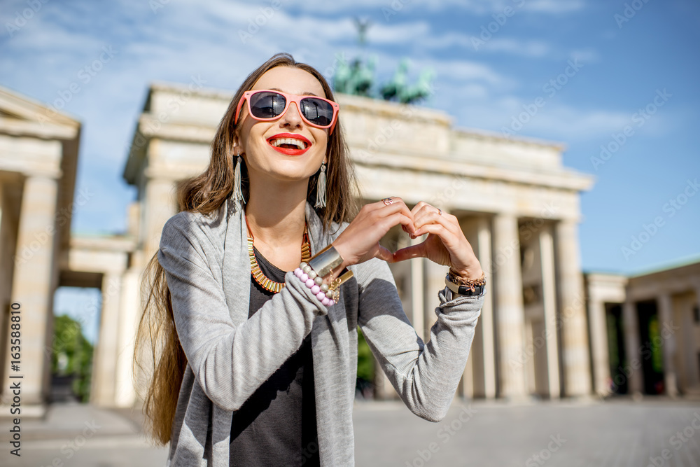 Fototapeta premium Young woman tourist making heart shape with hands in front of the famous Brandenburg gates in Berlin