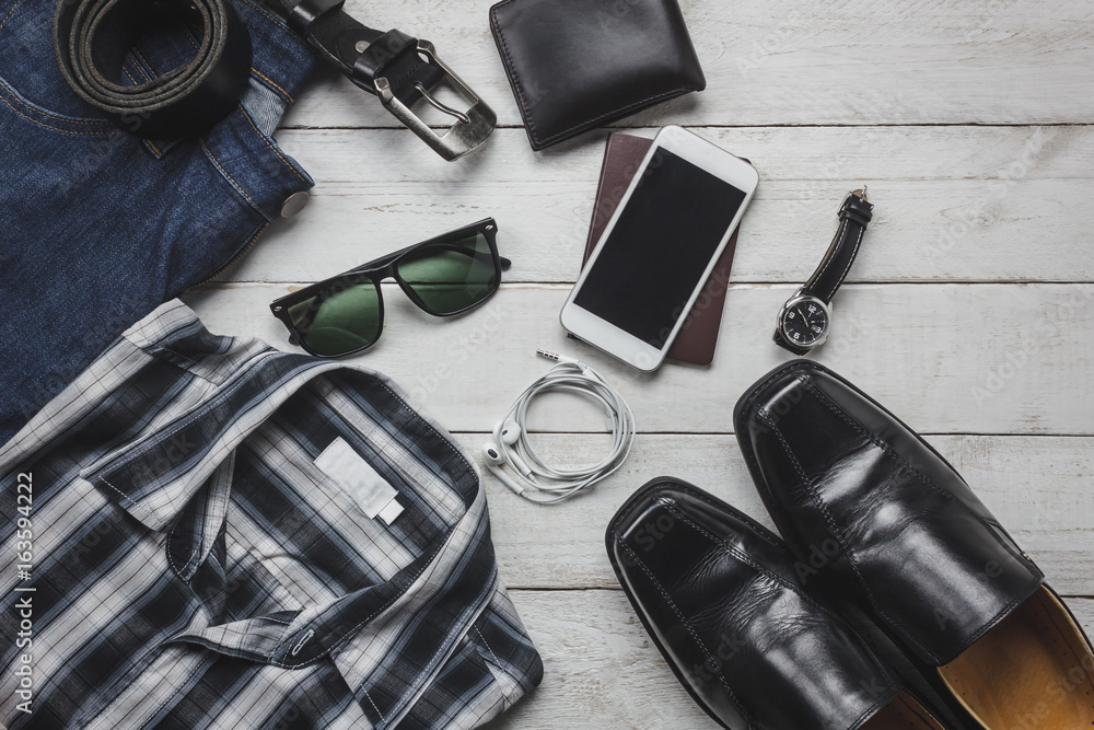 Top view accessoires to travel with man clothing concept.essential items for trip are shirt,jean,mobile phone on wooden background.watch,sunglasses and shoes on wood table.flat lay.copy space. Top view accessoires to travel with man clothing concept.essential items for trip are shirt,jean,mobile phone on wooden background.watch,sunglasses and shoes on wood table.flat lay.copy space.