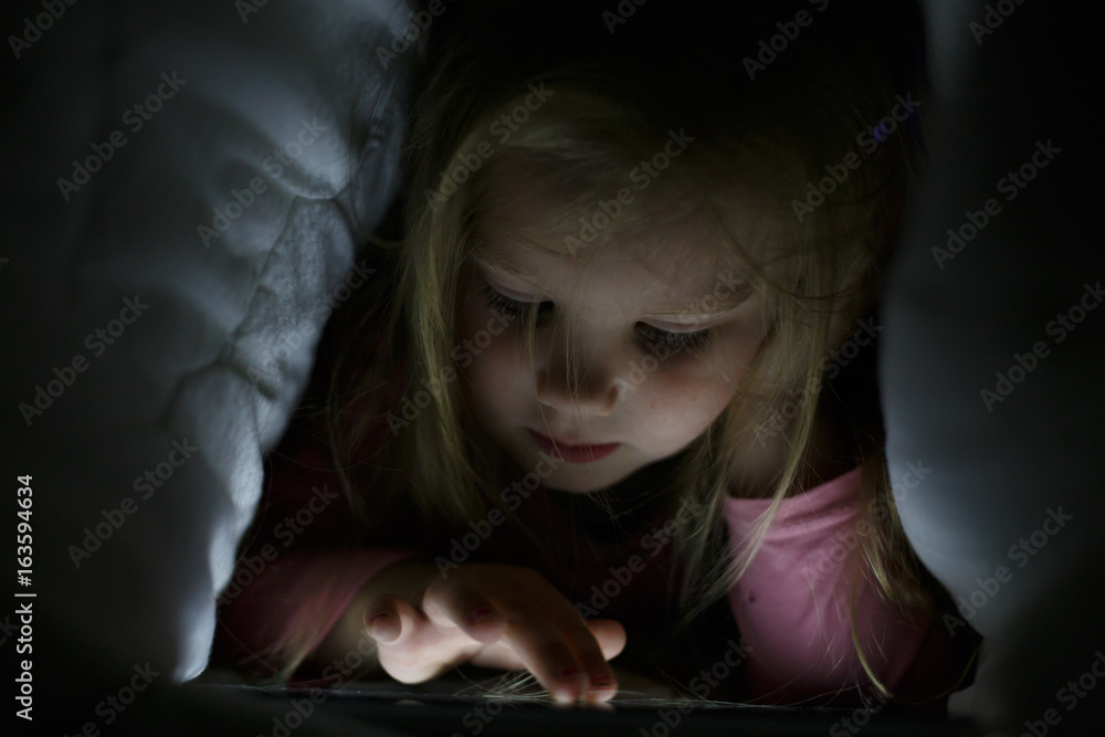 Little girl is playing on the tablet in secret from her parents at night under the blanket Little girl is playing on the tablet in secret from her parents at night under the blanket