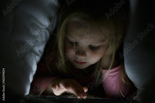 Little girl is playing on the tablet in secret from her parents at night under the blanket