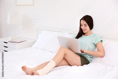 Beautiful young woman with laptop and bandaged leg sitting on bed at home
