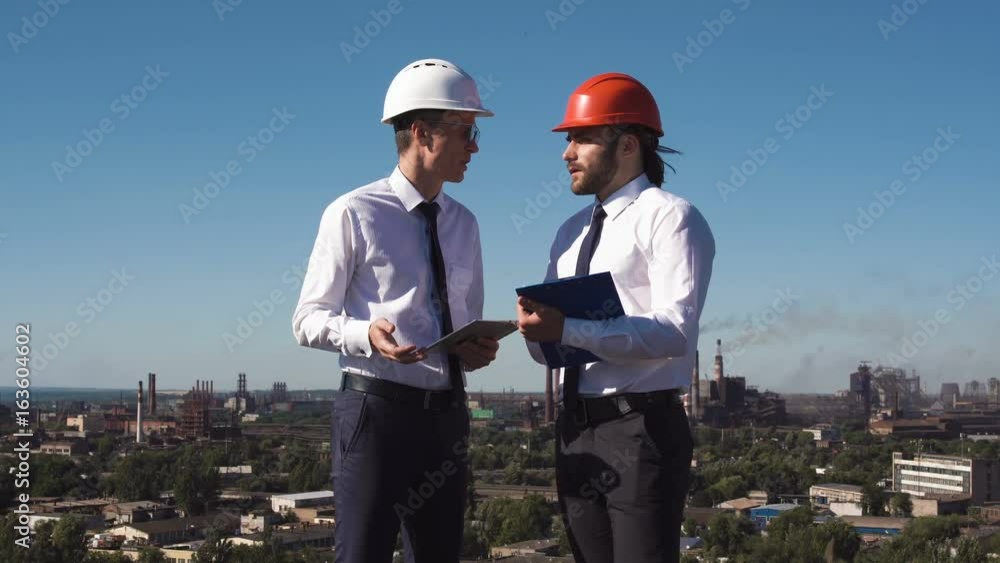 Businessmen in hardhats standing and discussing against the background of industrial Businessmen in hardhats standing and discussing against the background of industrial