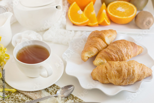 Continental breakfast with gold french croissants fruits and cup of tea on white table in a Morning light. Breakfast concept