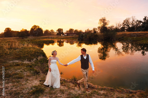 The bride and groom walk in the evening against the background of the lake in the red sunset. Overall plan