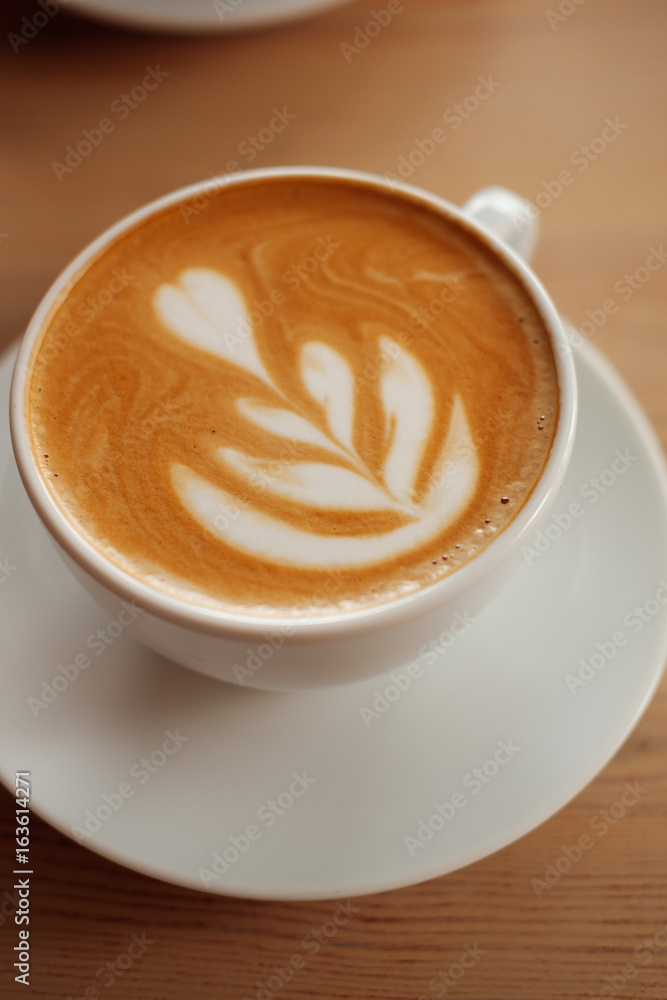 White cup of fragrant cappuccino stand on a wooden table. Coffee with milk on the table, front view. White cup of fragrant cappuccino stand on a wooden table. Coffee with milk on the table, front view.