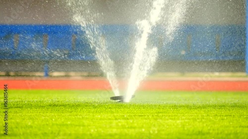 Watering the football field  by underground sprinklers. Lawn grass on the football field in hot summer, blue tribune seats  in background