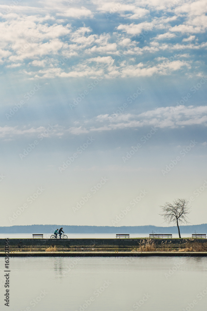 Couple of people riding a tandem bicycle in the distance near lake. Landscape with cloudy sky and reflection in the water. A lot of copy space for text.