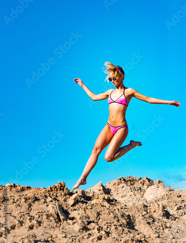 Photo of beautiful young model jumping, in the air, on pile of sand and blue sky background. Vertical view