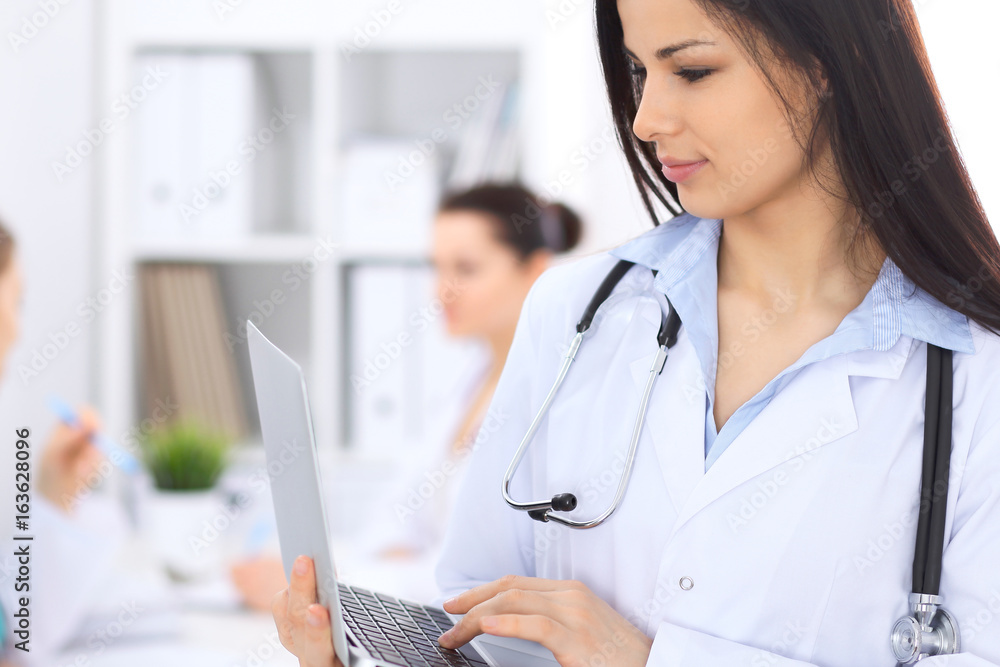 Brunette female doctor on the background of colleagues talking to each other in hospital. Physician is ready to help patients. Medicine and health care concept Brunette female doctor on the background of colleagues talking to each other in hospital. Physician is ready to help patients. Medicine and health care concept