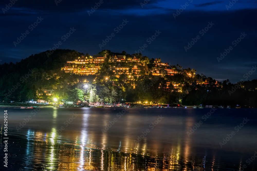 Romantic outdoor photography on a night beach of Phuket with many lights from houses on a hill, light reflection on a sea shore and traditional thai boats floating in water of BangTao beach, Thailand