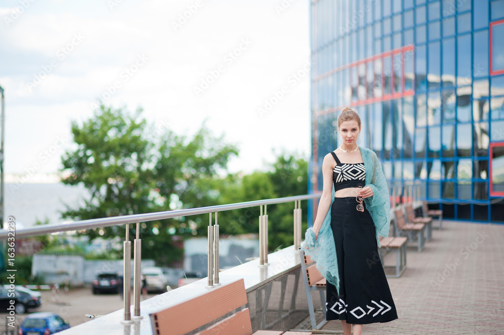 A beautiful girl in a black top and a long skirt on a walk against a blue mirror building. A girl with a tender blue, turquoise stole. Portrait of a girl with glasses in a cityscape. Flying knitted mo