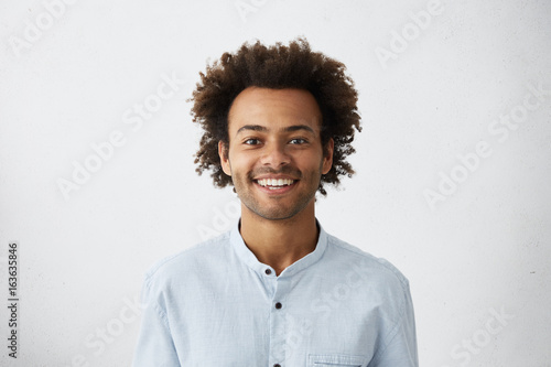 Positive guy with African hairstyle and dark skin wearing elegant white shirt looking cheerfully into camera isolated over white background. Young dark-skinned male with smile dressed elegantly