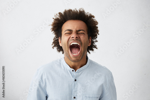 Portrait of dark-skinned African guy with curly hair screaming with wide opened mouth closing his eyes in despair having unhappy expression. Frustrated male shouting loudly having aggression © wayhome.studio 