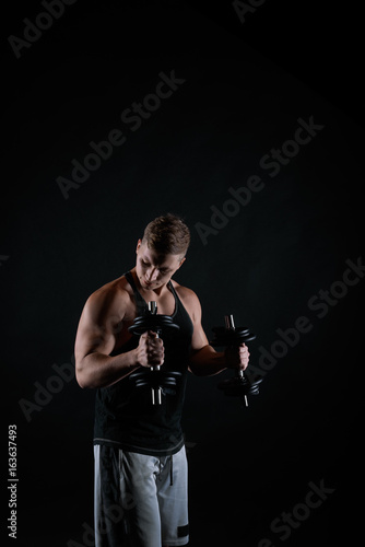 Dark concept of hard training on black background. Athletic brutal man working with dumbbells. Bodybuilder trainer in t-shirt and grey shorts. Muscular guy making exercises.