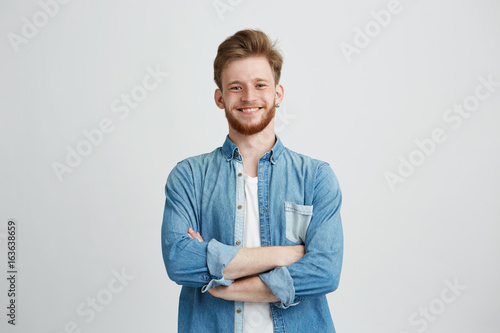 Portrait of young handsome man in jean shirt smiling looking at camera with crossed arms over white background.
