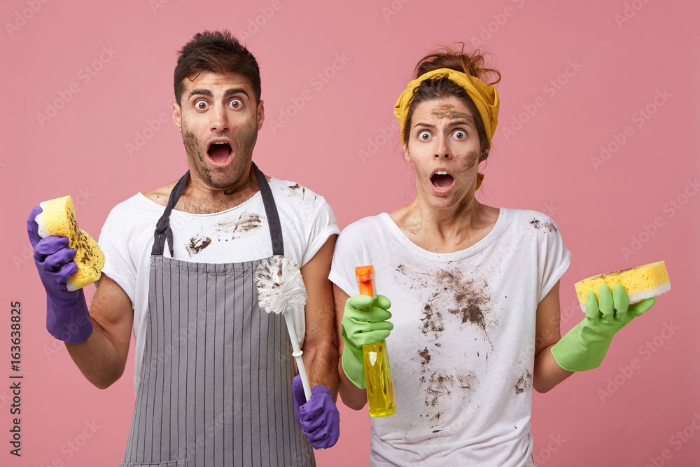 Φωτογραφία Handsome male wearing apron standing near his wife holding cleaning equipment having shocked look