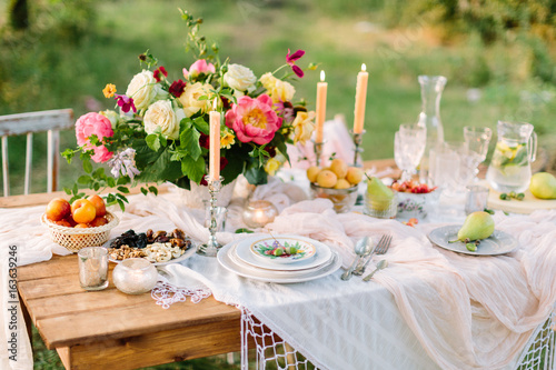 women's day, wedding, celebration, romance, picnic, nature concept - gorgeous table setting with snow white tablecloth, dishes, clear wine-glasses, silver candleholders and various fruits