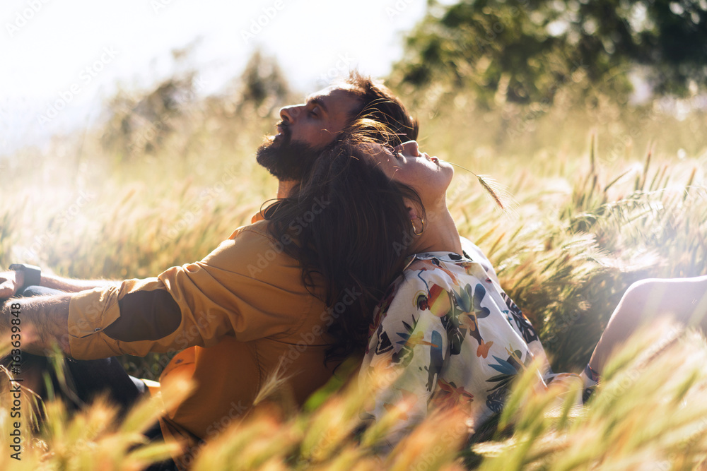 A happy beautiful couple in love is sitting amidst the meadow on a warm sunny day. Romantic photo of a man and a woman sitting back-to-back on a grass. Weekend picnic dating outdoors. - obrazy, fototapety, plakaty 