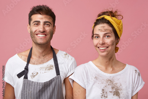 Studio portrait of smiling man and woman in white T-shirts being dirty after cleaning their apartment isolated over pink background. Houseowners with glad experssions looking directly into camera photo