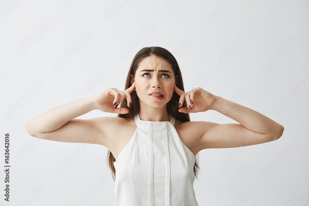 Displeased beautiful brunette girl closing ears with fingers from noise biting lip over white background. Displeased beautiful brunette girl closing ears with fingers from noise biting lip over white background.