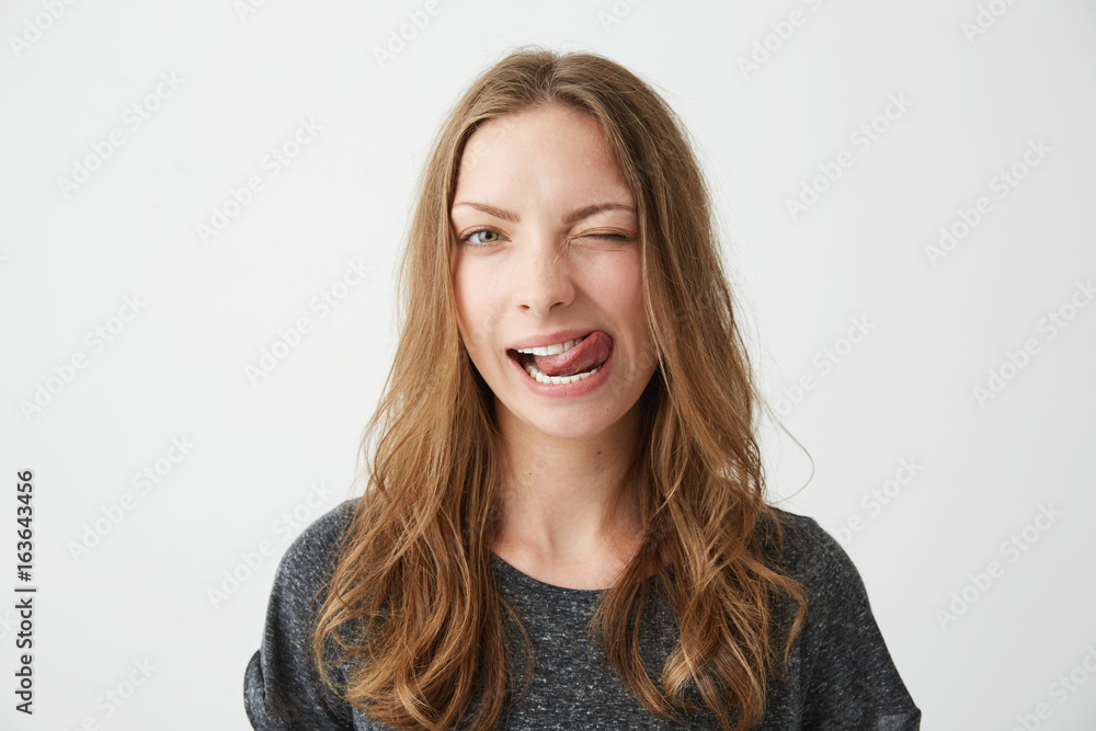Portrait of young positive pretty girl looking at camera winking showing tongue over white background. Portrait of young positive pretty girl looking at camera winking showing tongue over white background.