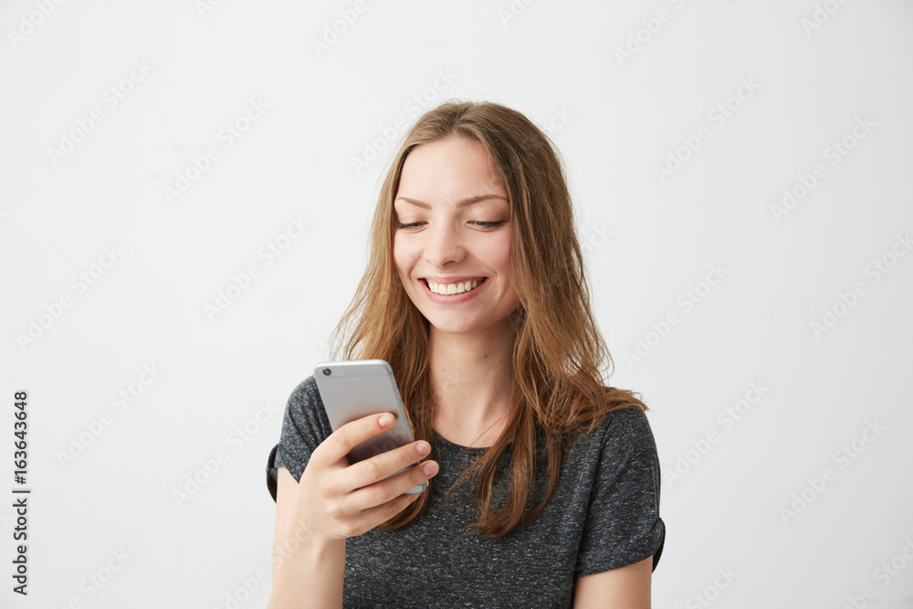 Happy cheerful girl smiling looking at phone screen typing text message over white background. Happy cheerful girl smiling looking at phone screen typing text message over white background.