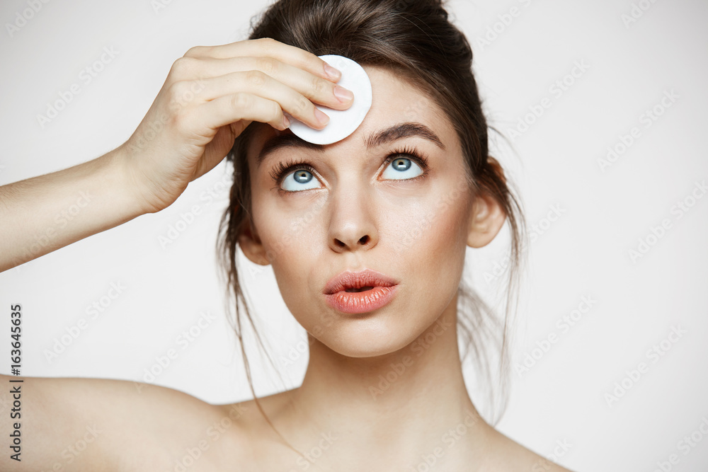 Fototapeta premium Cute beautiful natural brunette girl cleaning face with cotton sponge smiling over white background. Cosmetology and spa.