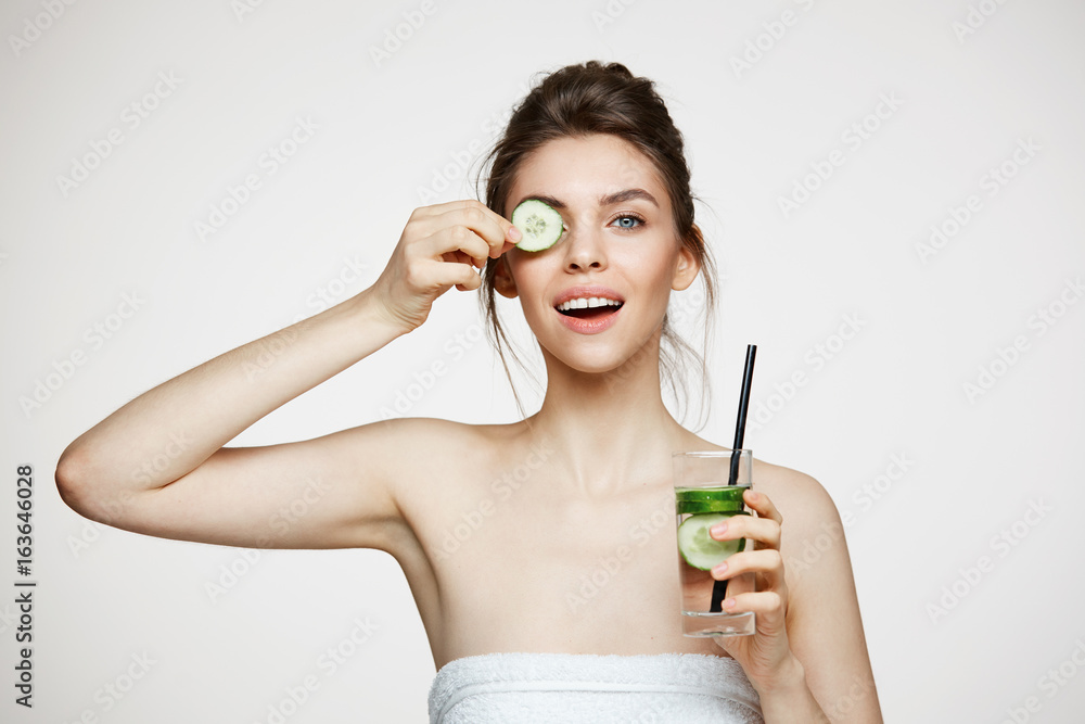 Positive young brunette girl smiling looking at camera holding cucumber slice holding glass of water over white background. Beauty and health. Positive young brunette girl smiling looking at camera holding cucumber slice holding glass of water over white background. Beauty and health.