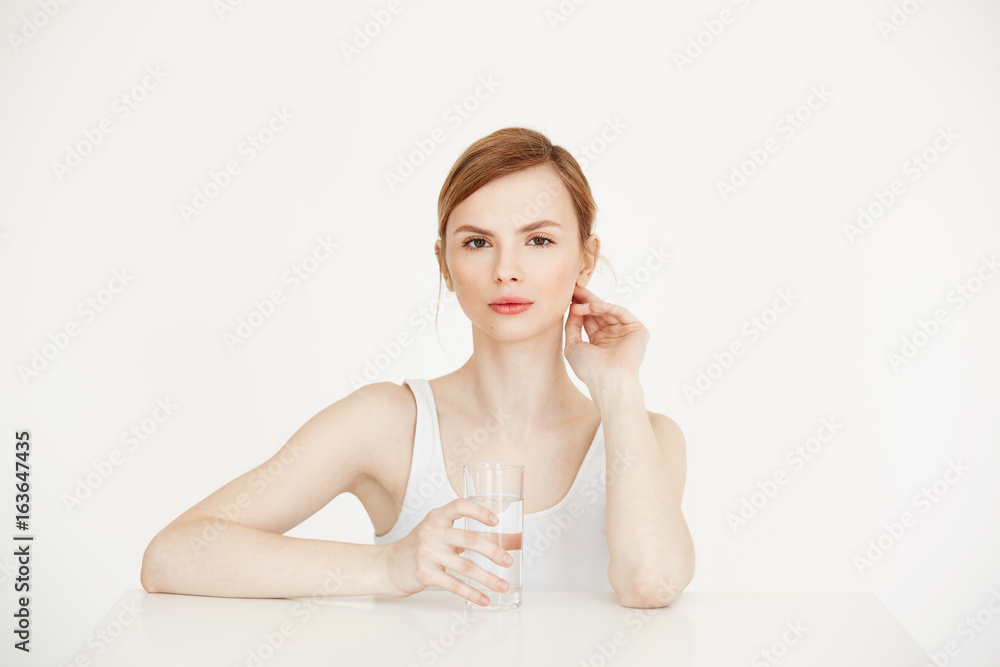 Young beautiful girl with clean fresh skin holding glass with water sitting at table over white background. Health and beauty lifestyle. Young beautiful girl with clean fresh skin holding glass with water sitting at table over white background. Health and beauty lifestyle.