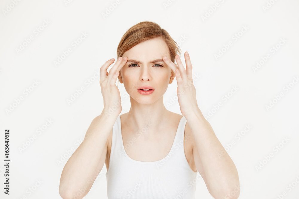Young beautiful girl touching skin looking at camera sitting at table over white background. Cosmetology skincare and beauty. Young beautiful girl touching skin looking at camera sitting at table over white background. Cosmetology skincare and beauty.