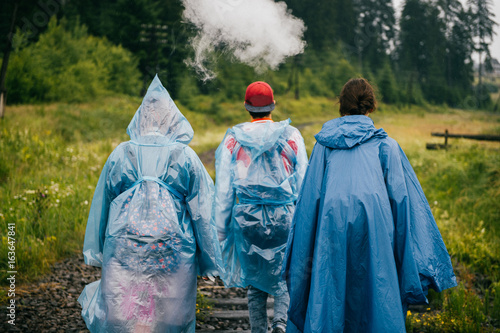 Smoking man and two women. Rebel travelers in raincoats walking in danger zone on railroad outdoor at narure. Occurrence on road. Warning. Accident. Company of friends on vacation. Tourist clothes.