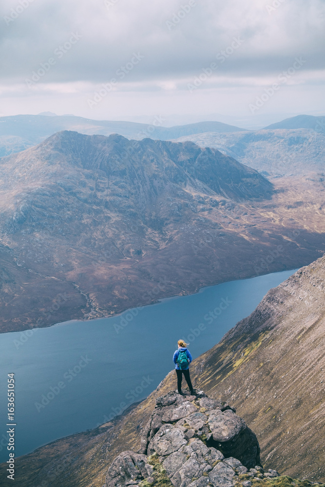 Small figure of female hiker standing at the edge of a cliff adoring the view. An Taellach ridge, Highlands of Scotland, UK