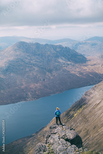 Small figure of female hiker standing at the edge of a cliff adoring the view. An Taellach ridge, Highlands of Scotland, UK