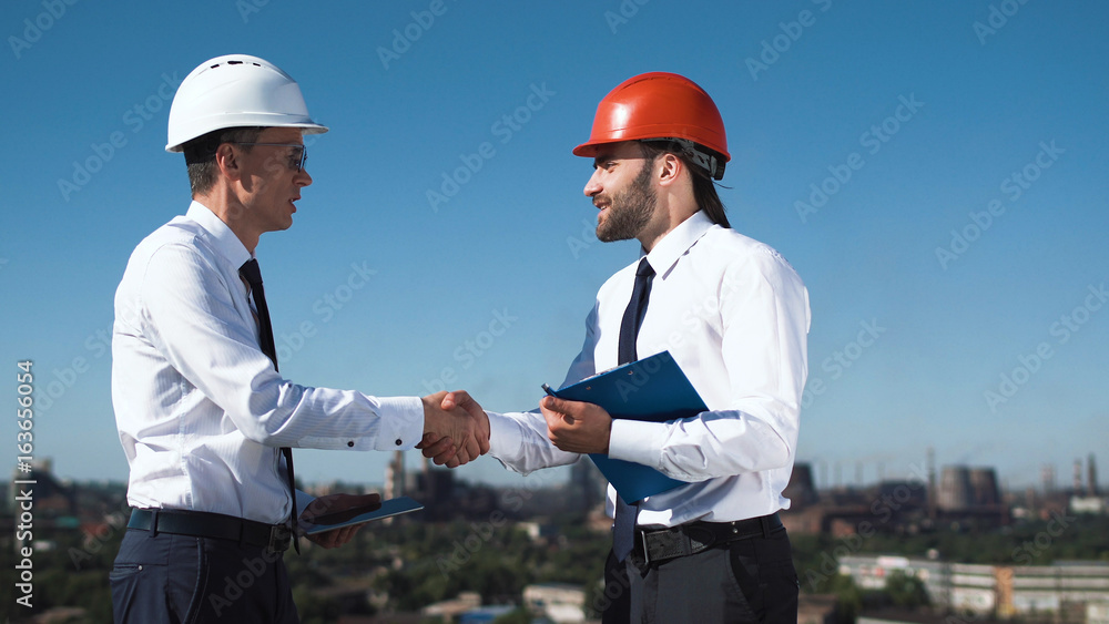 Businessmen in hardhats standing and discussing and shaking hands making a deal with factory view at background Businessmen in hardhats standing and discussing and shaking hands making a deal with factory view at background
