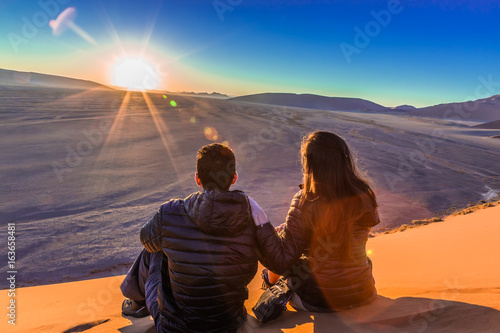 Couple on sand dune in desert during sunrise.  Sossusvlei, Namib Naukluft National Park, Namibia. Real Lens Flare Effect