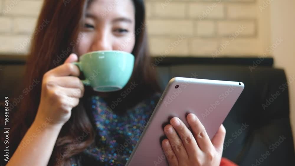 A beautiful Asian woman with smiley face holding and using tablet pc while drinking hot coffee in modern cafe A beautiful Asian woman with smiley face holding and using tablet pc while drinking hot coffee in modern cafe