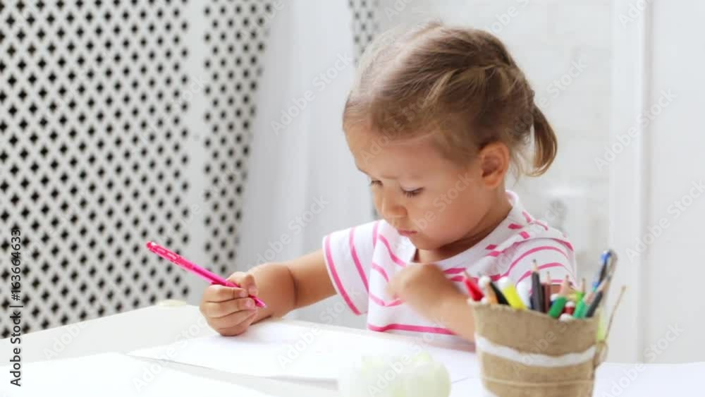Cute preschool girl sitting by the white table focused on drawing something. Cute preschool girl sitting by the white table focused on drawing something.