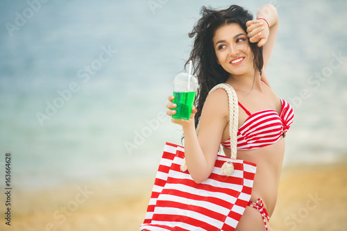 Beautiful woman at the beach having a tan in the red swimsuit with cold freshing drink and bag at the seaside in the summer heat