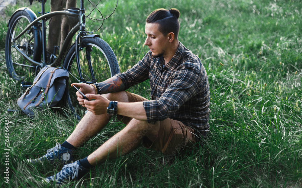 Young Male Cyclist Sits On Summer Meadow Near Bicycle, Holding At Tablet Recreation Resting Travel Destination Concept