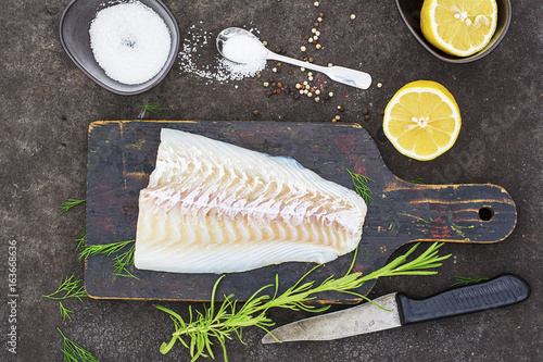 Raw cod before cooking on a black chopping Board with herbs and sea salt on a dark background. Top view photo