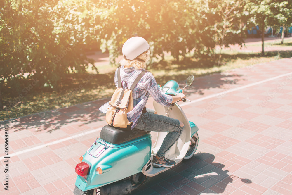 Fototapeta premium Młoda wesoła dziewczyna jazdy skuterem w mieście. Portret młodej i stylowej kobiety z motorowerem.