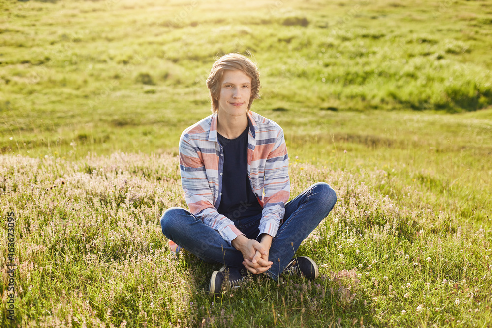Candid shot of handsome teenager boy sitting crossed legs at green grass of field looking directly into camera with pleased expression keeping his hands together while having good rest outdoors Candid shot of handsome teenager boy sitting crossed legs at green grass of field looking directly into camera with pleased expression keeping his hands together while having good rest outdoors