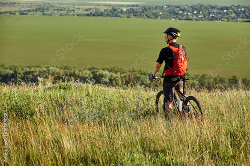 Rear view of the sportsman with his mountain bike stands on the meadow and looking away in the countryside.