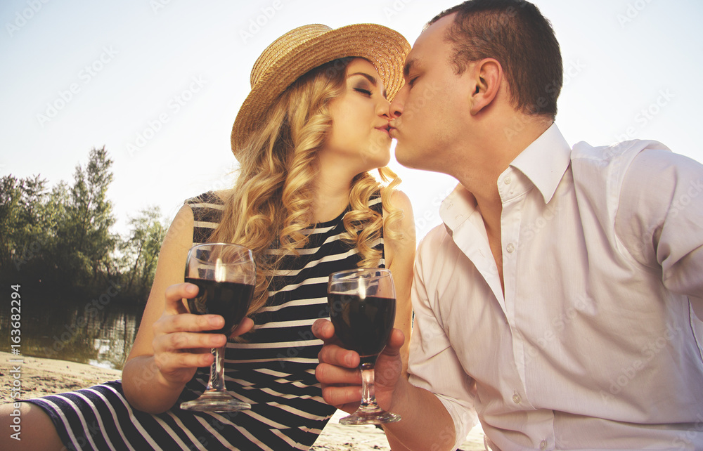 Love to kiss her lips. Portrait of man in casual wear kissing his young  beautiful woman in dress and keeping glasses with wine in their hands while  having picnic on beach. Stock