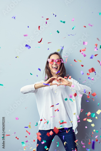 Laughing young woman dressed in heart shaped glasses making hear gesture at camera with confetti around her