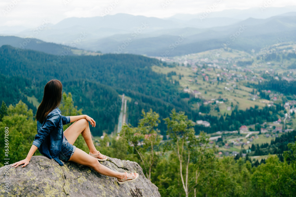 Obraz premium Lonely young traveler girl siiting on stone at top of mountain. Discovering new country. Beautiful nature landscape from high altitude. Summer trip on weekend vacation. Edge of world. Jeans clothes.