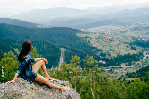 Lonely young traveler girl siiting on stone at top of mountain. Discovering new country. Beautiful nature landscape from high altitude. Summer trip on weekend vacation. Edge of world. Jeans clothes.
