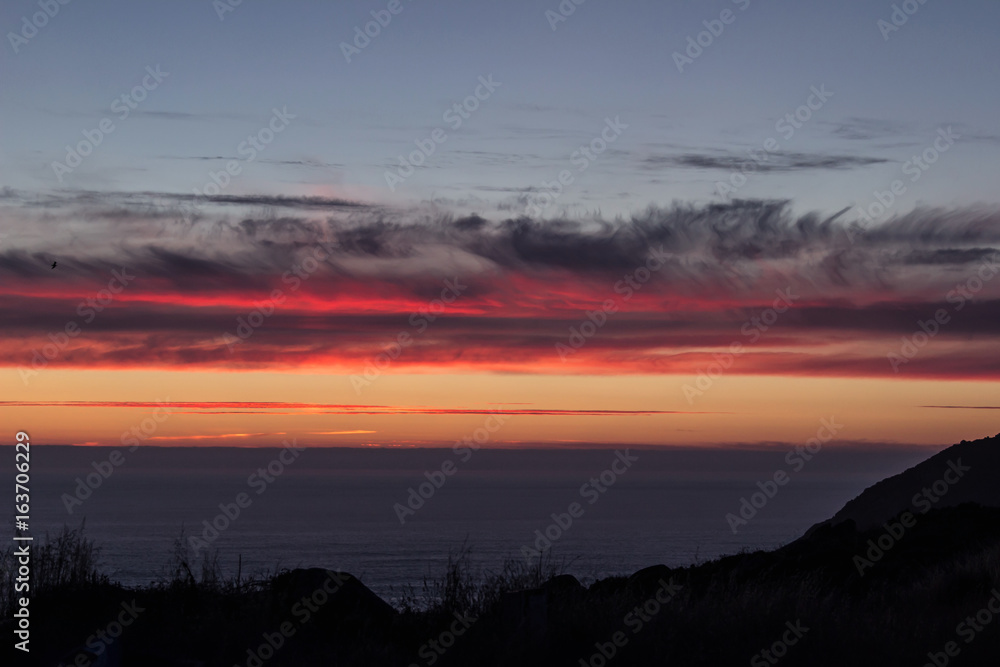 Fototapeta premium Shot of the Atlantic Ocean, with red sunset clouds, and the silhouette of the cliffs in Finisterre, Galicia, Spain