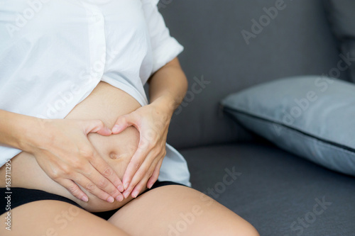 Pregnant asian woman holding gesture heart symbol shape belly with on sofa in home. Maternity concept.